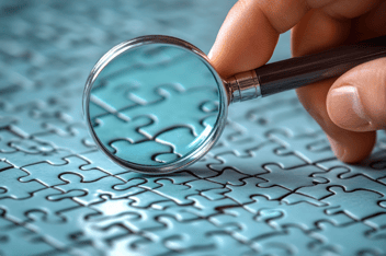 Close-up of a hand holding a magnifying glass over a blue jigsaw puzzle, looking at some interlinked pieces.